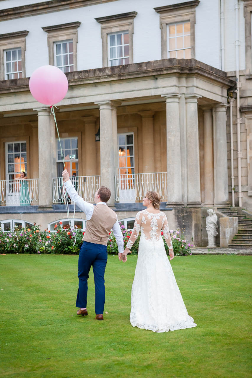 couple walking away from camera holding hands with groom holding large pink balloon