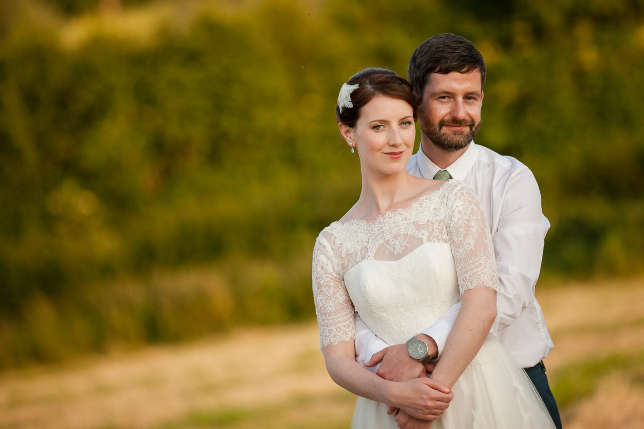 groom standing behind bride with arms wrapped around her waist, both looking into camera