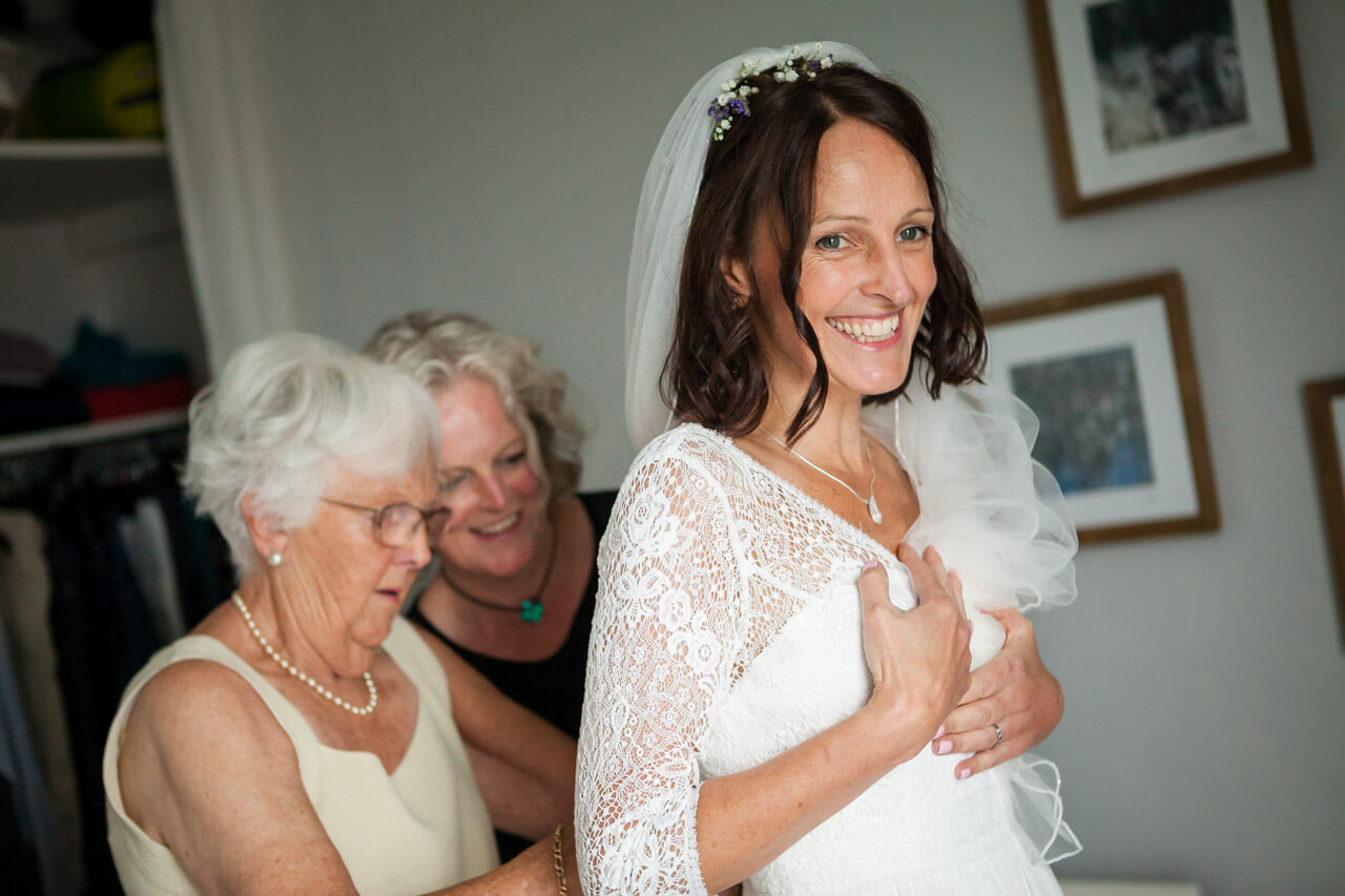 bride smiling while mother and sister doing up back of wedding dress