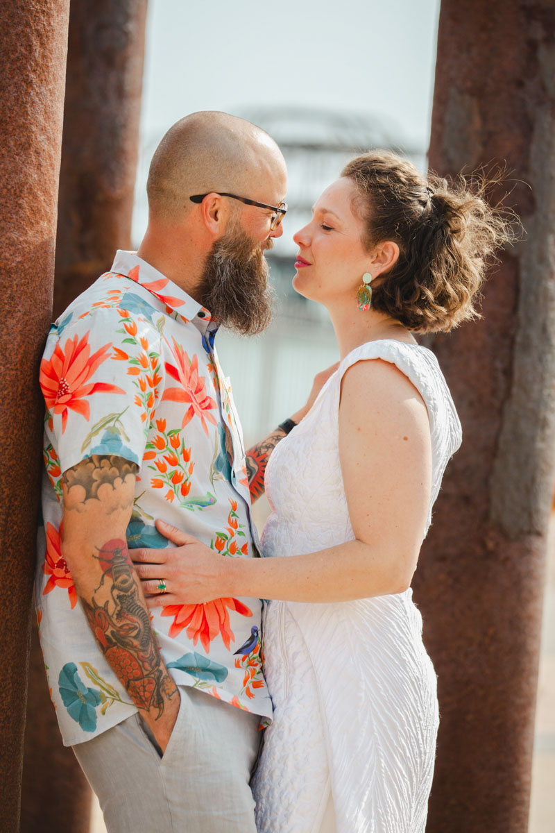 bride puts arms around groom leaning against pillar west pier