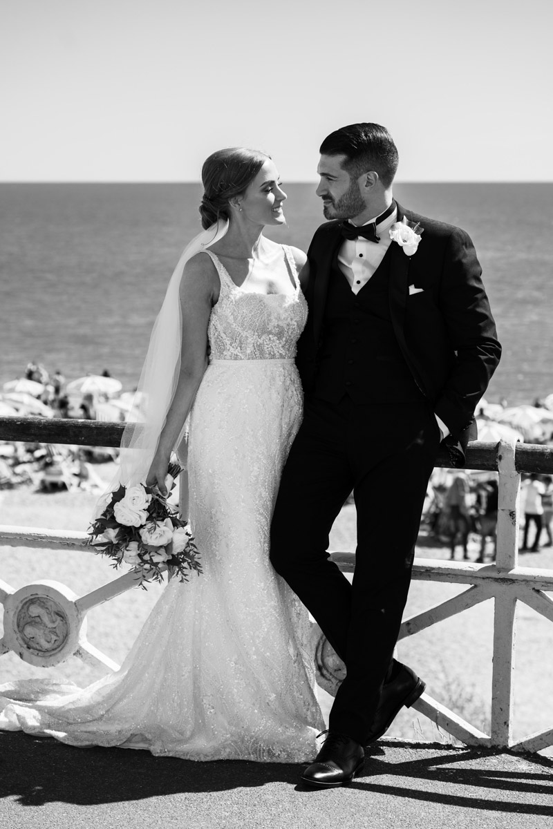 bride and groom standing on sea front looking into each others eyes. Groom with his hands in his pocket