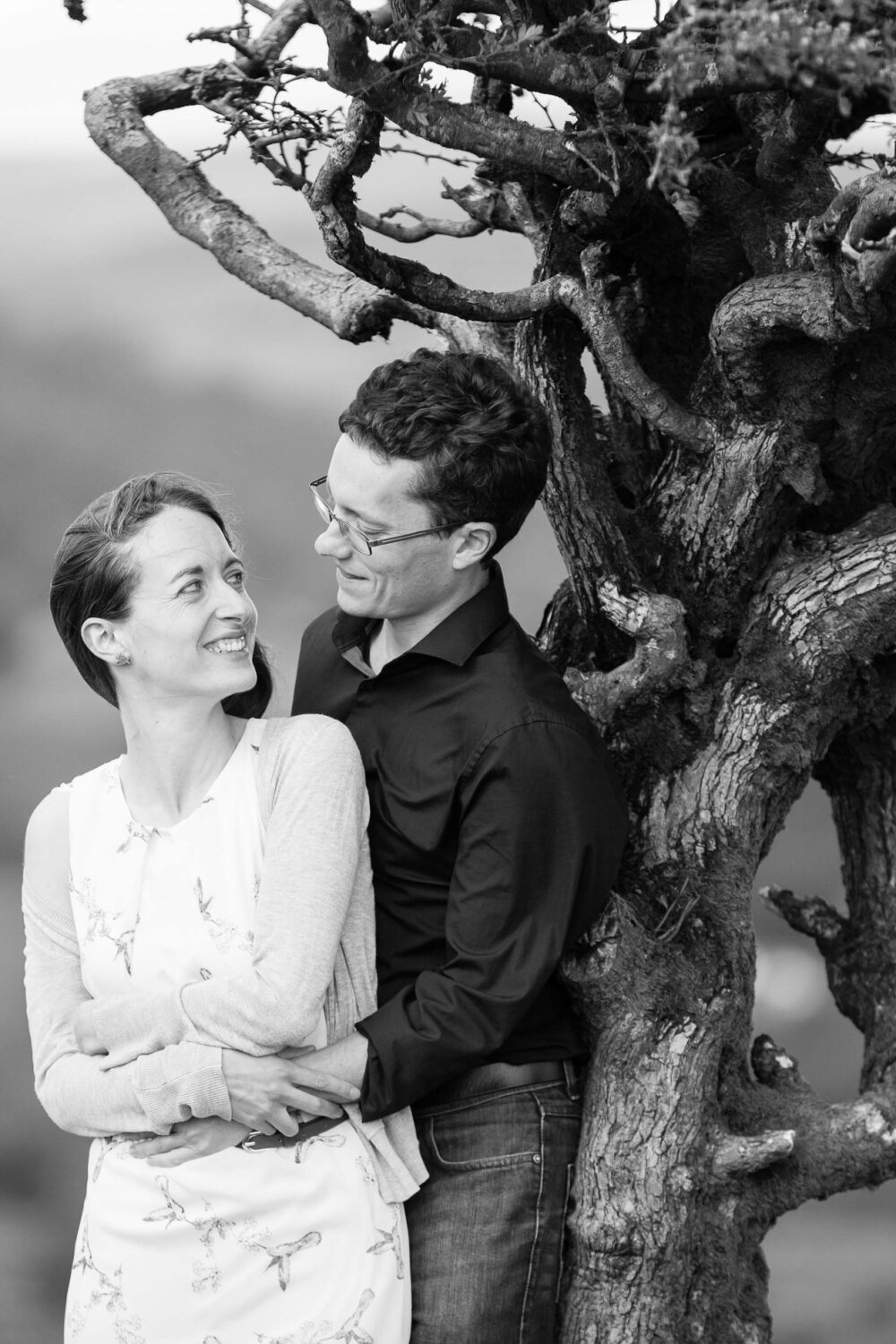 man leans against tree looking towards his fiancée who is looking over her should back at him