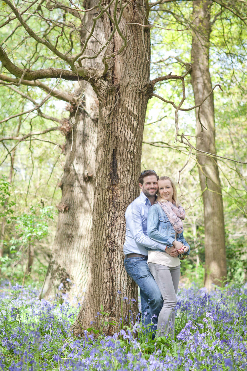 in woods, man and woman lean against tree and each other looking towards camera