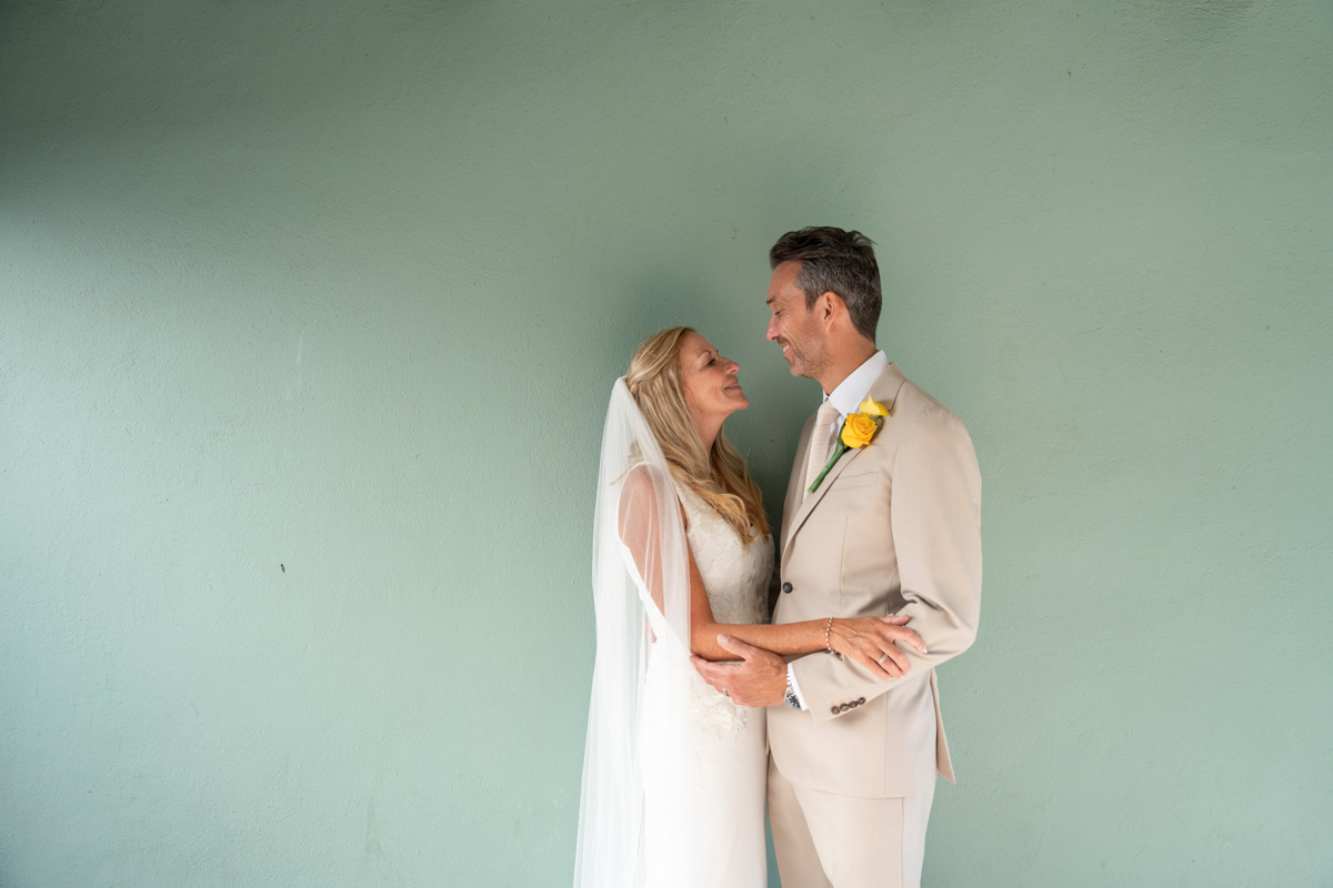 bride and groom look into each others eyes standing facing each other against plain green wall background