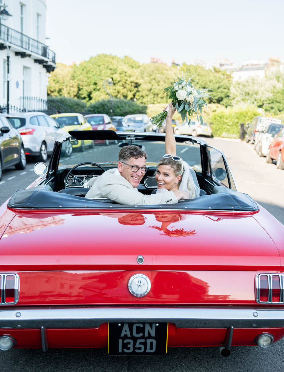 bride and groom looking backwards in mustang car holding bouquet in the air