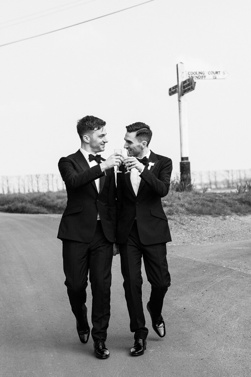 two grooms walking in their tuxedos clinking champagne glasses as they walk
