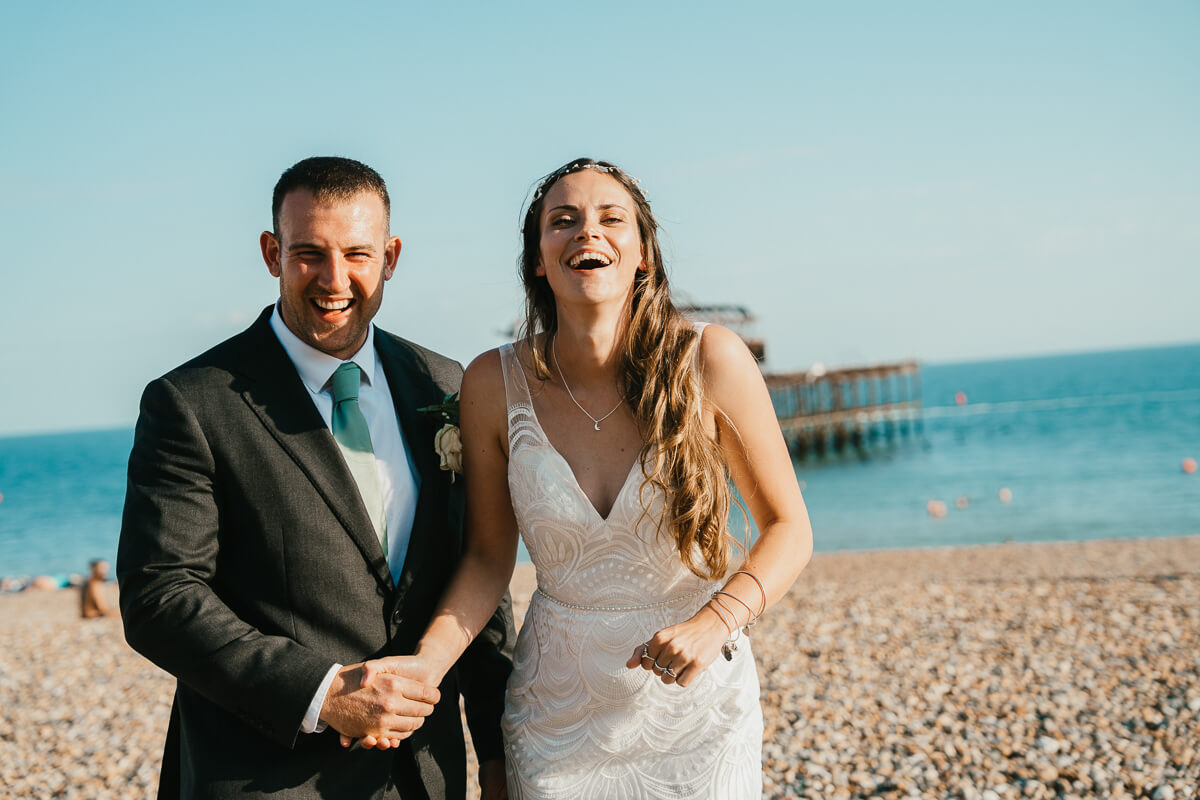 bride and groom laughing towards camera on Brighton beach with sea in background