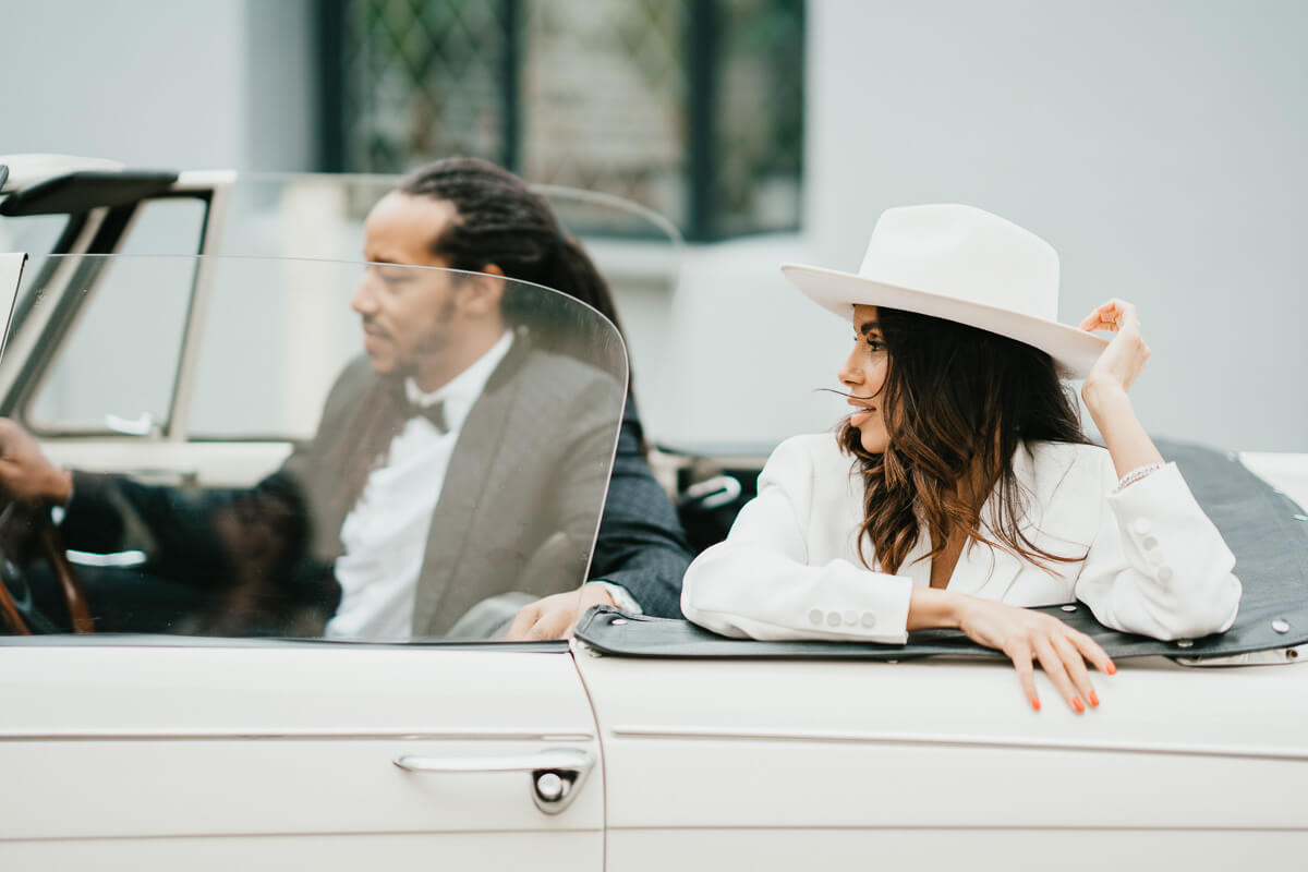 groom sitting hold drivers wheel in classic car with bride in the back arm sitting on side of car