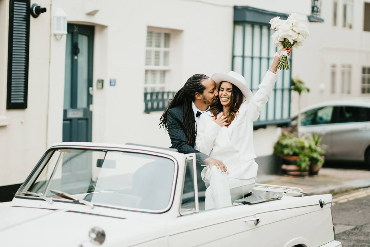 bride and groom sitting on the back of vintage car with groom sitting brides face and bride smiling holding up her bouquet