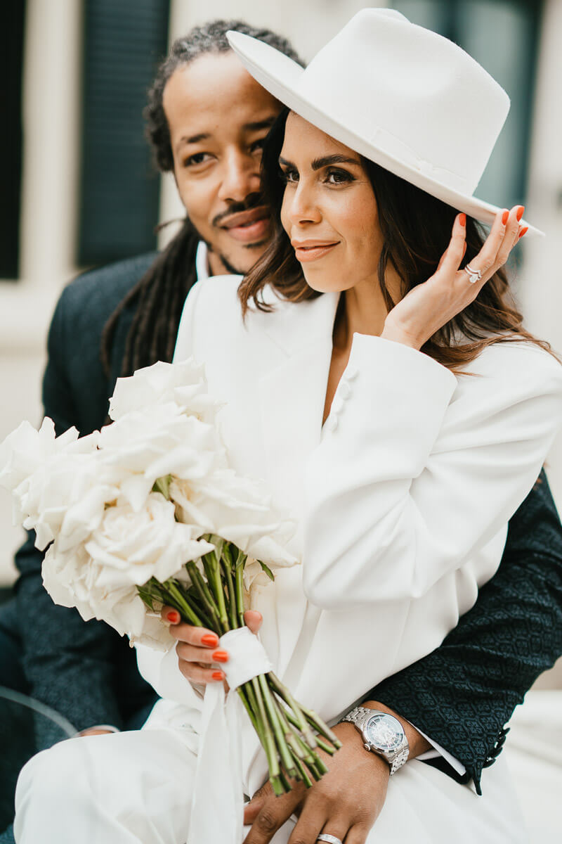 bride holding her hat and flowers smiling while groom tucks arms around bride both looking away from the camera