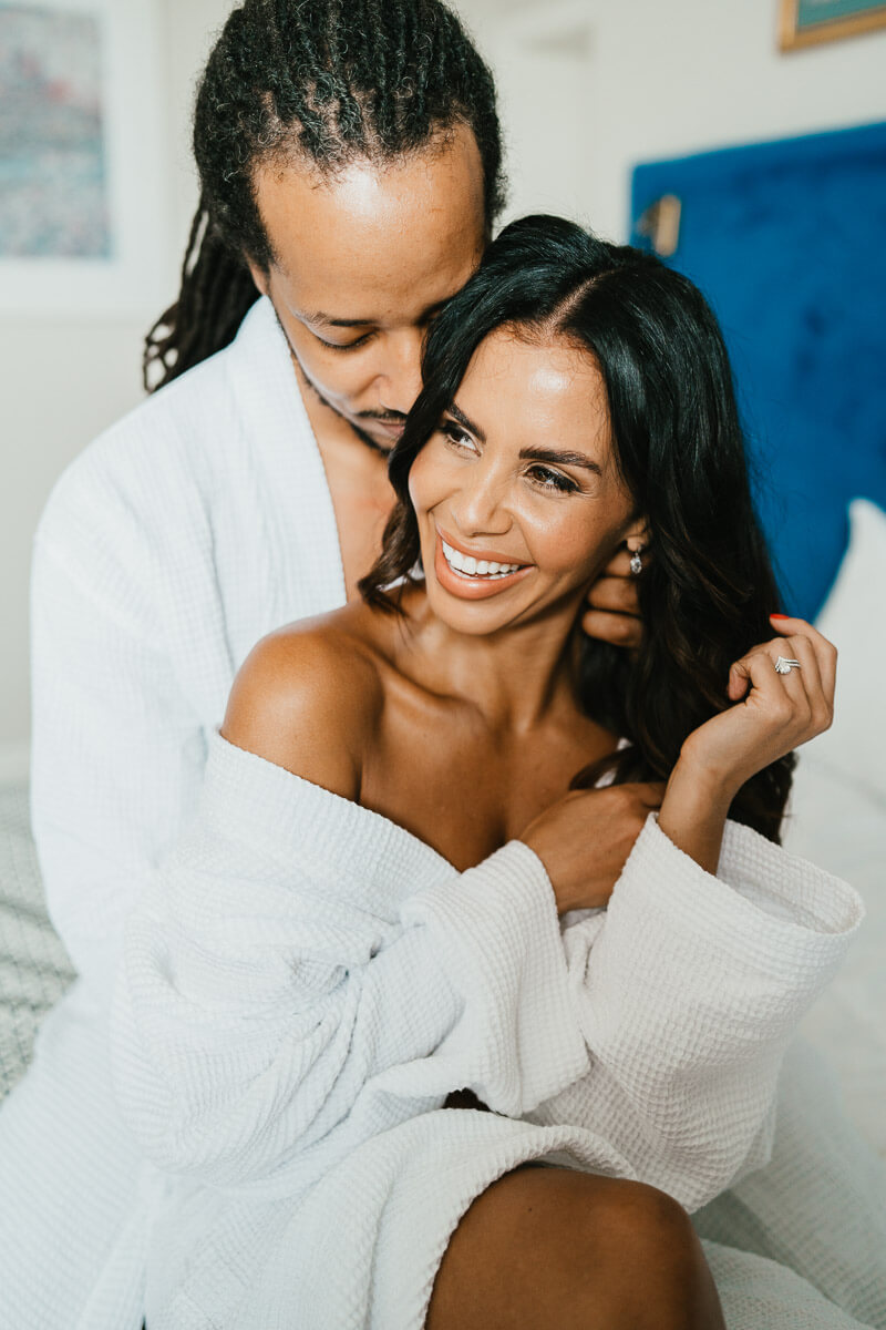 bride smiling sitting on bed in dressing gown with groom behind her tucked into her ear