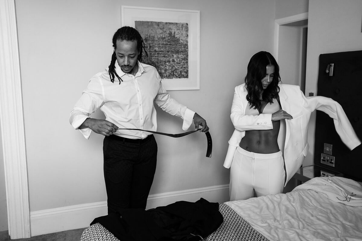 bride and groom getting ready in hotel room. bride putting on jacket groom putting on tie
