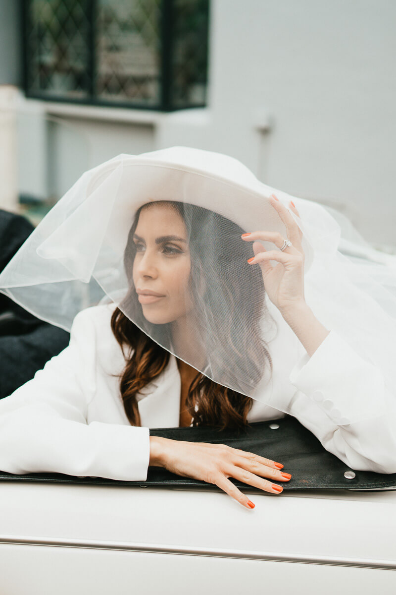 bride holding hat with netting veil covering her face while sitting in back of car