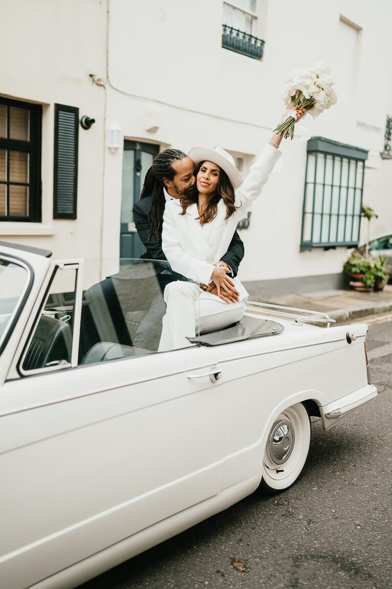 couple sitting on the back top of the car groom arms around bride kissing neck bride holding her flowers in the air