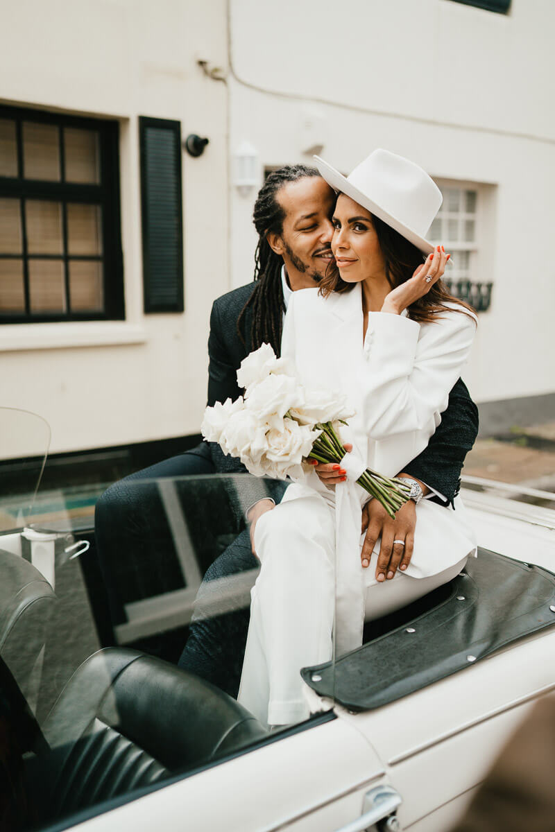 bride sitting crossed legged on back of car holding side of hat while groom sits slightly behind her touching her cheek