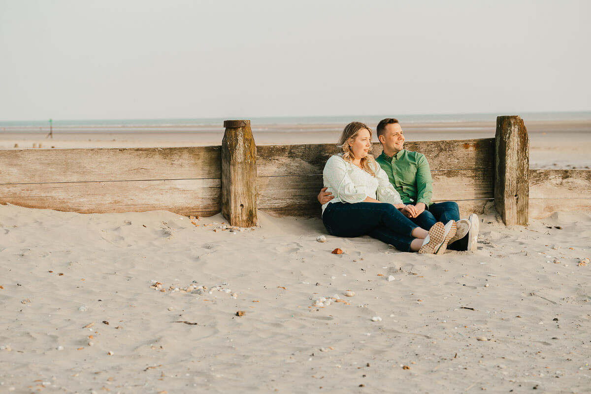 couple looking towards the sunset as they sit against wooden groin on beach