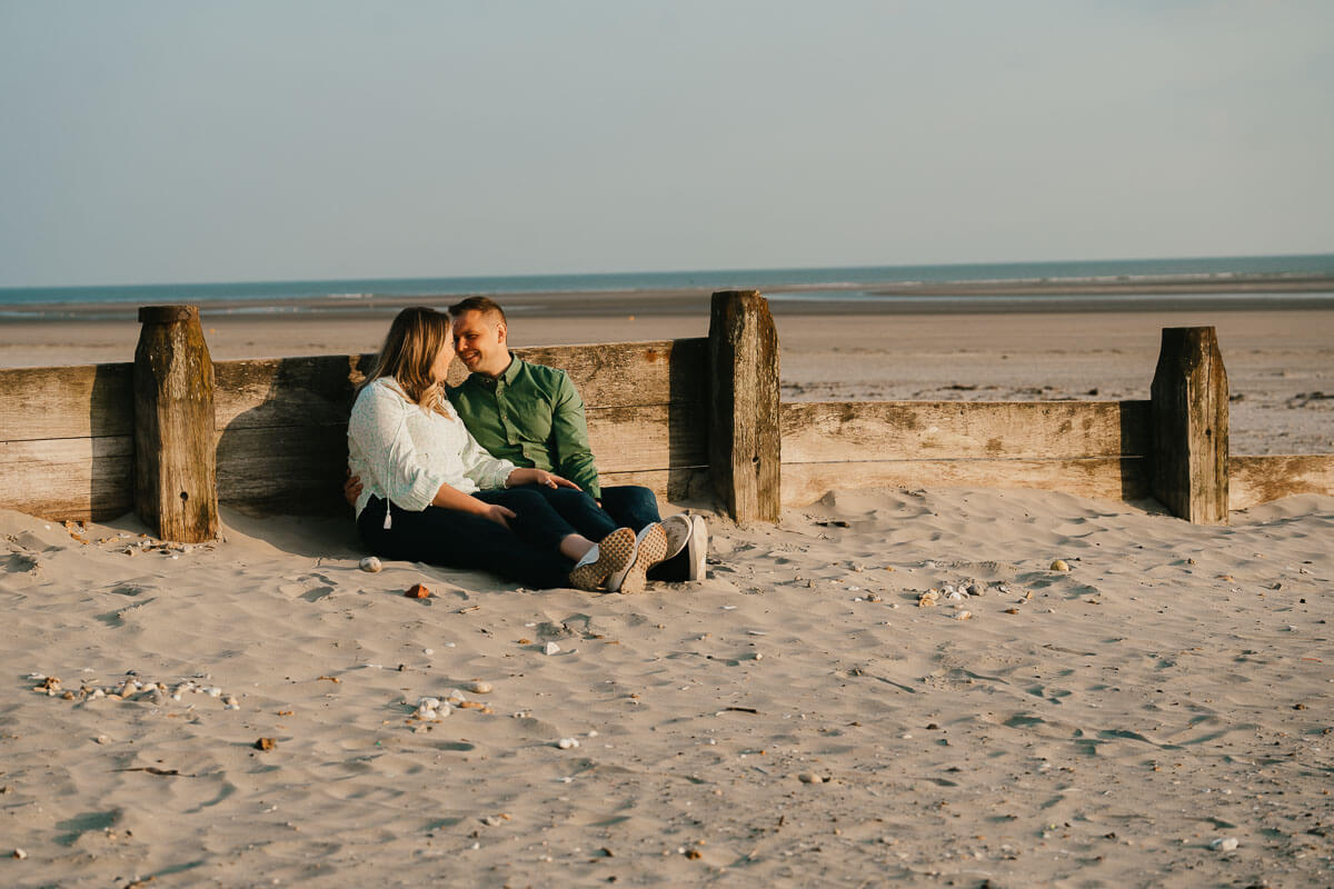 couple sitting against wooden beach groin looking at each other during golden hour