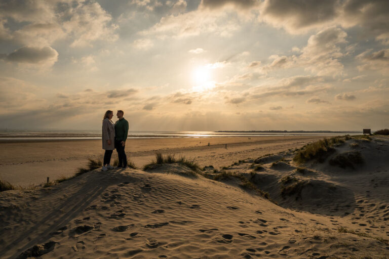 Couple standing on top of a sand dune with sun setting in the background