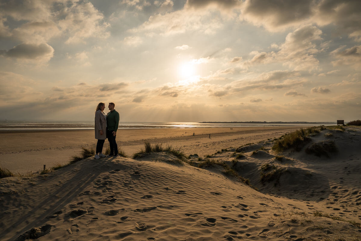 Couple standing on top of a sand dune with sun setting in the background