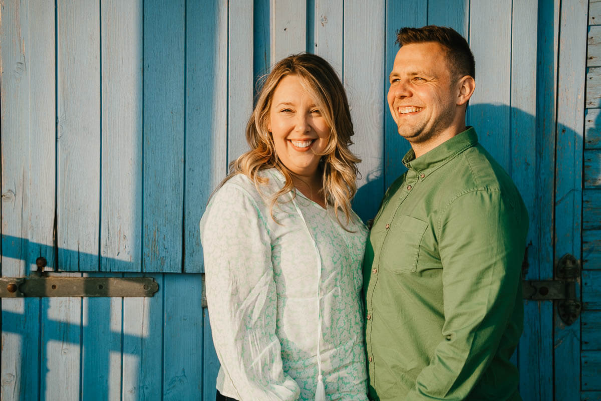 woman looking into the camera with her partner looking to the sunset standing infront of blue beach hut