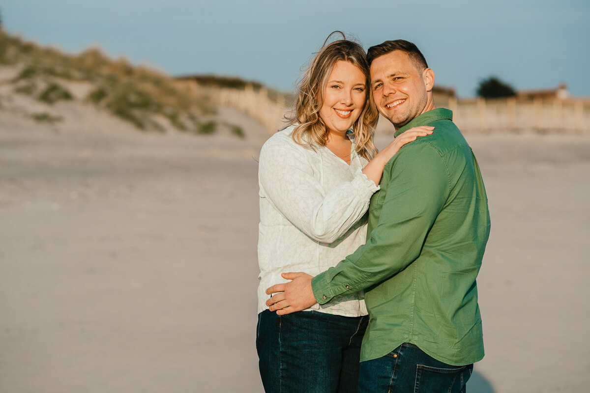 couple with arms around waist and shoulders looking into camera standing on beach