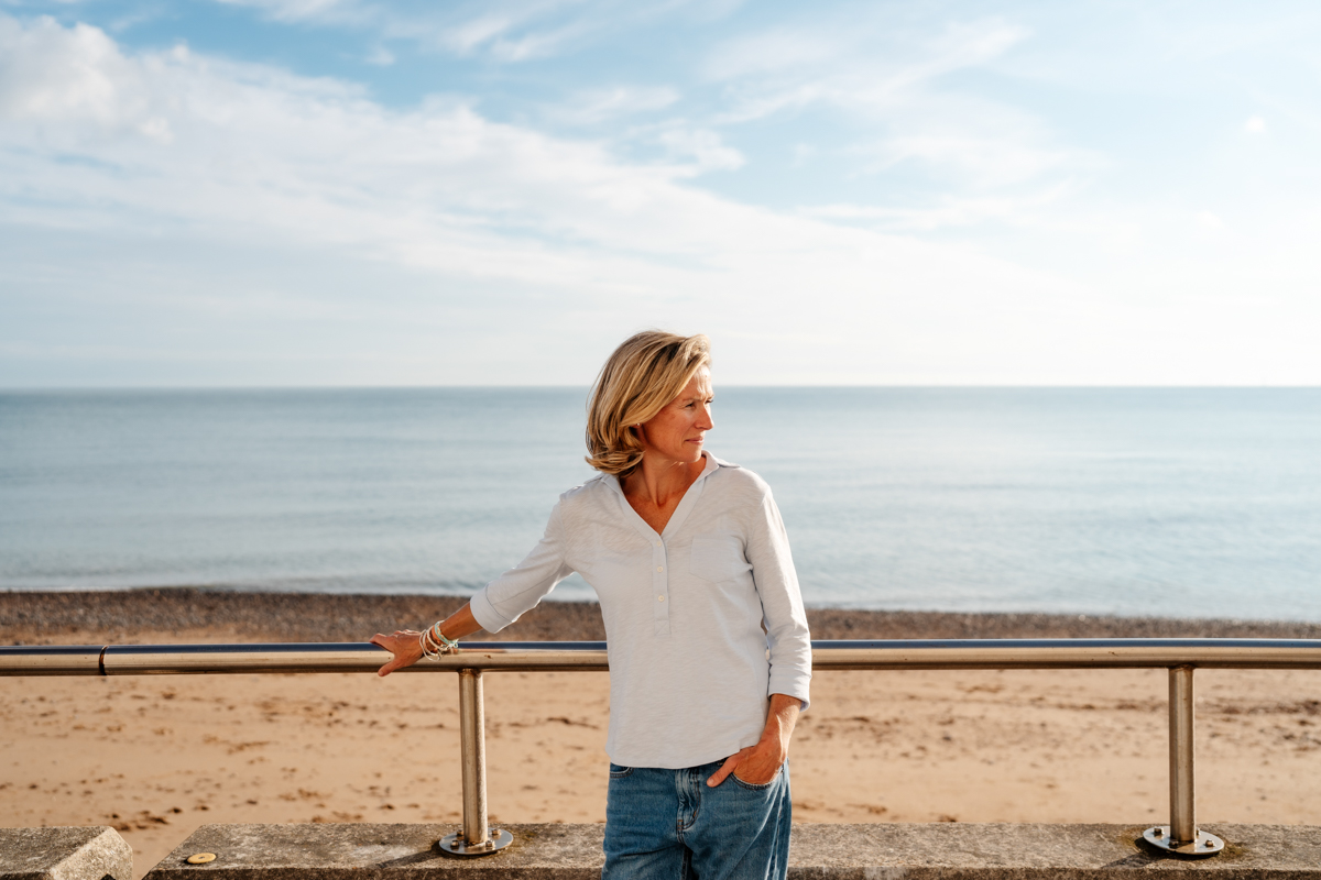 woman standing besides railings with pebble beach behind her looking out to sea