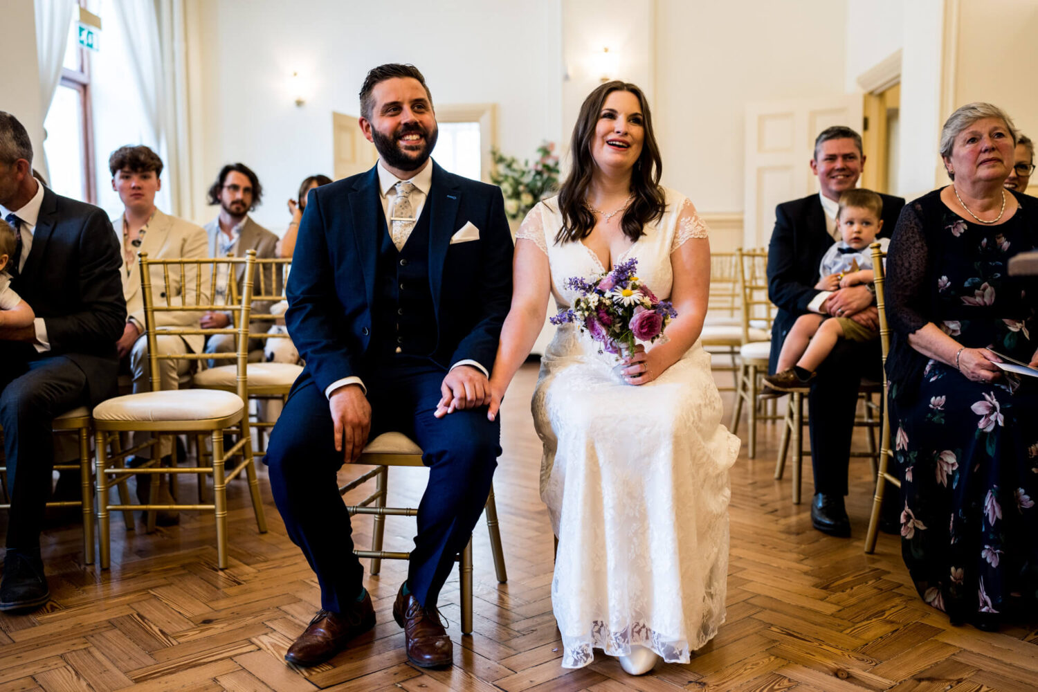 bride and groom sitting in chairs holding hands listening to registrar