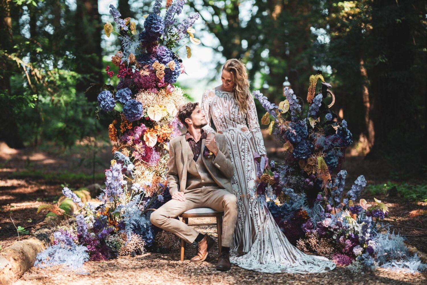 groom sitting on chair looking up at bride with hands linked. Amazing florals surrounding them in redwood forest
