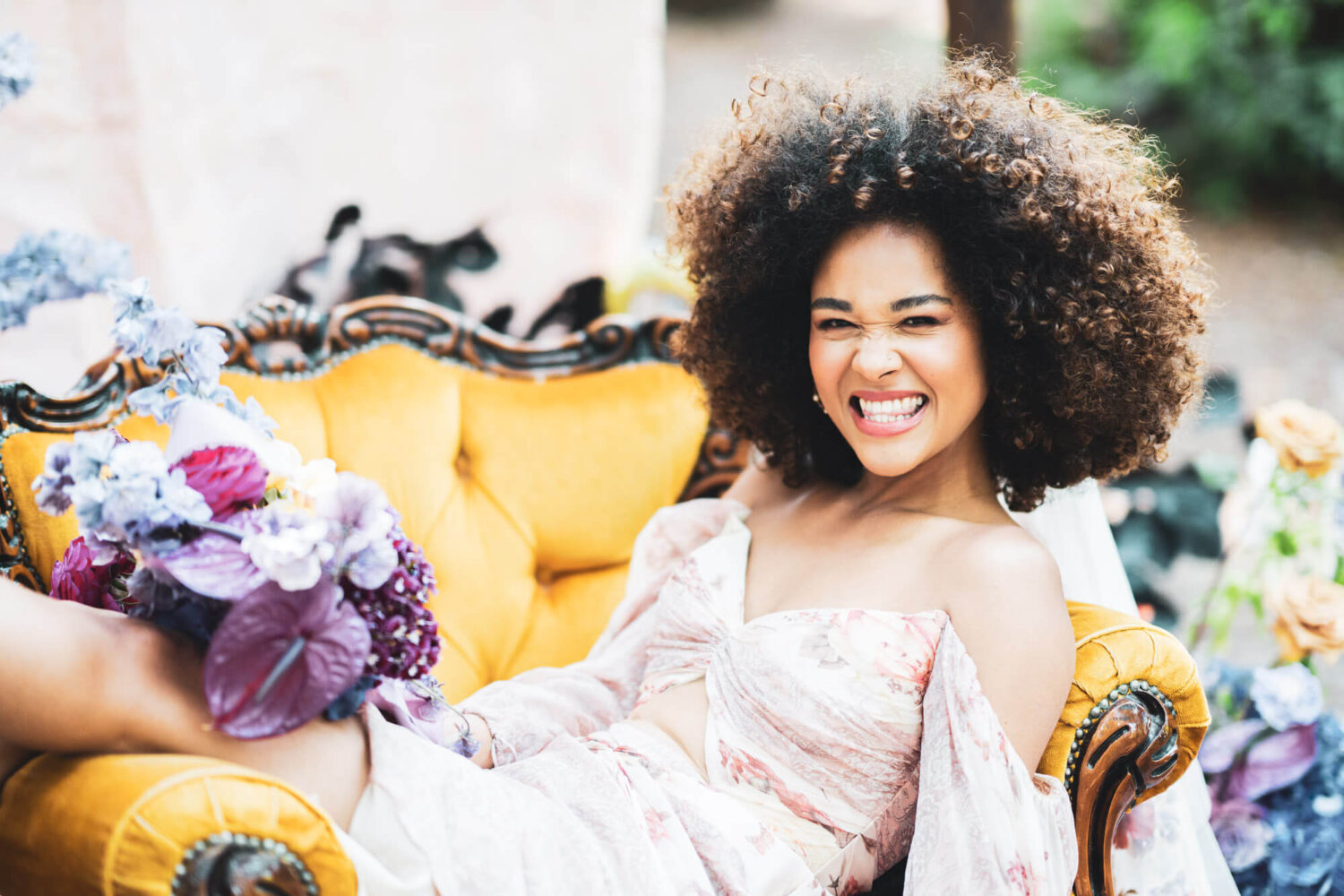 woman sitting in yellow chair with awesome floral colours around her cheeky smile into camera