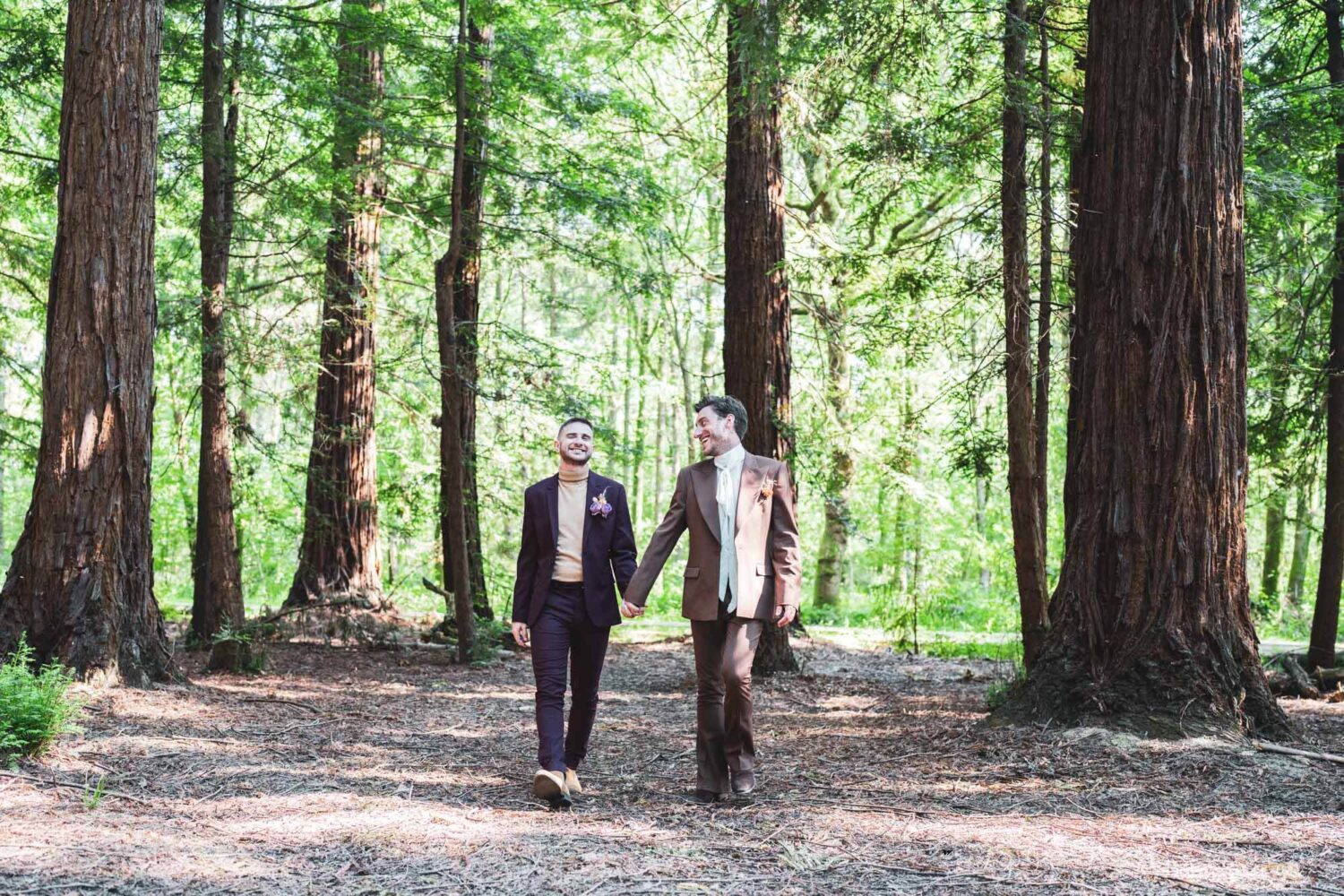 two grooms walking through redwoods tree forest holding hands looking at each other