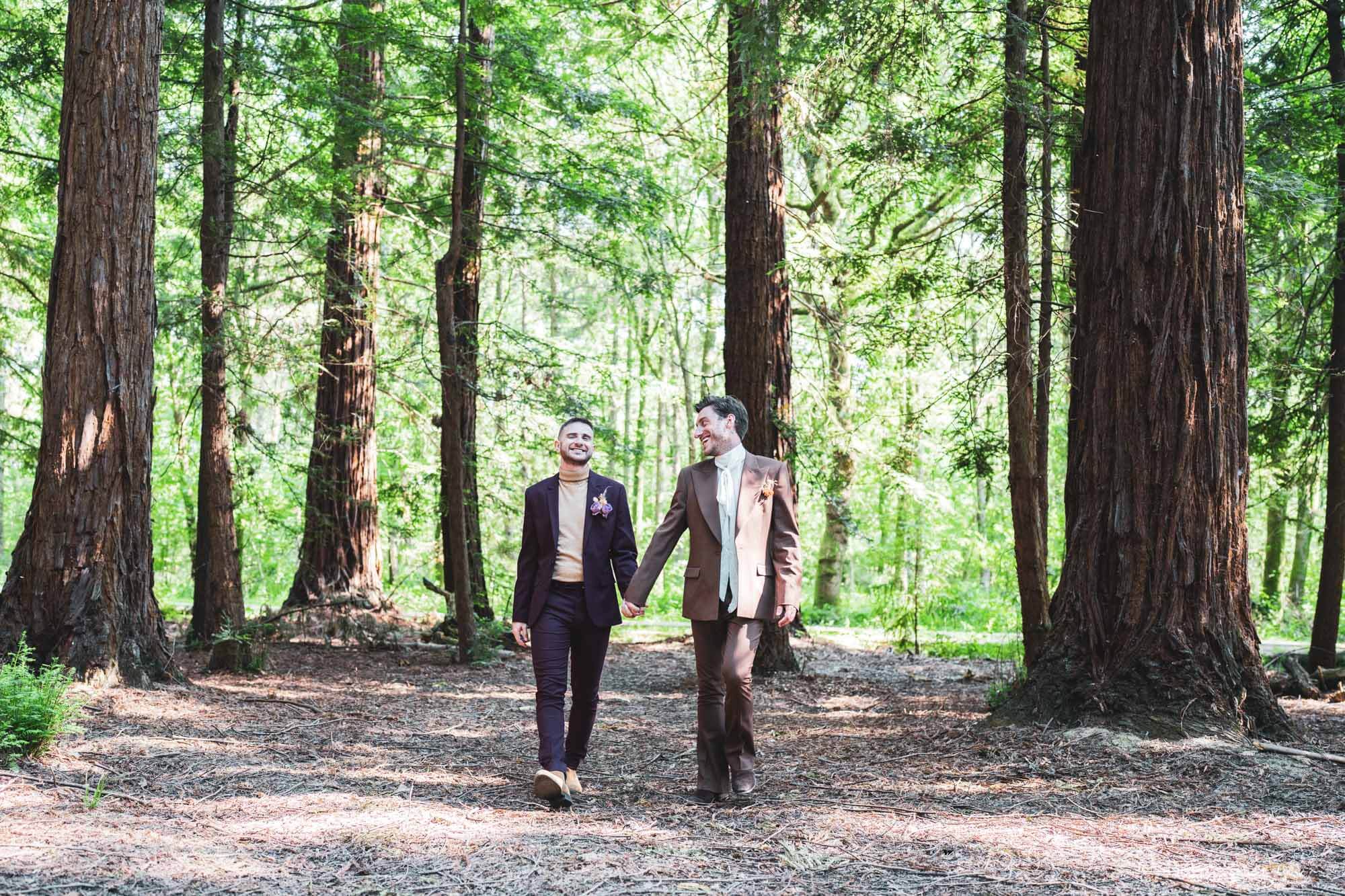 two grooms walking through redwoods tree forest holding hands looking at each other
