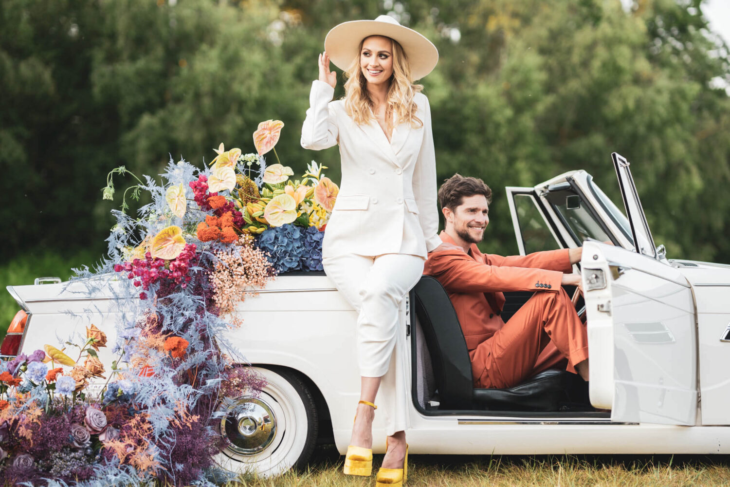 bride lean against vintage car with hand on wide rimmed hat while groom sits in the drivers seat