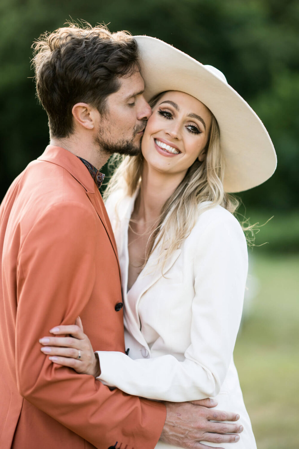 golden hour portrait of groom and bride with groom kissing side of bride's face and her looking into the camera