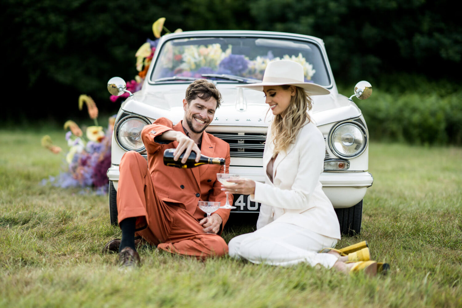 groom pours champagne into bride's glass sitting in front of vintage Triumph car