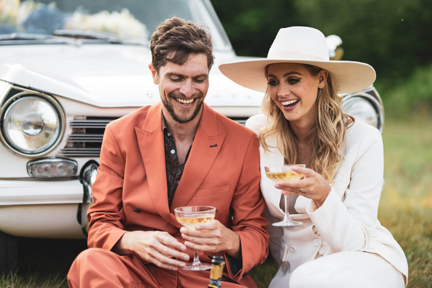 bride and groom enjoying a laugh while sitting on grass in front of Triumph car