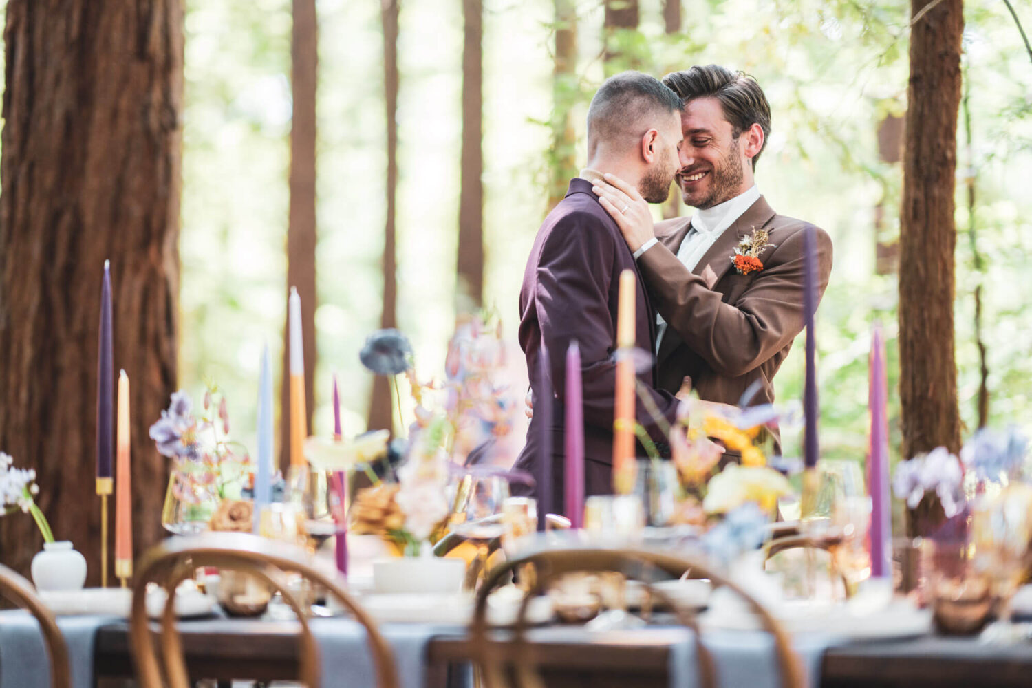 two grooms with heads together arms around each other with table set up in foreground