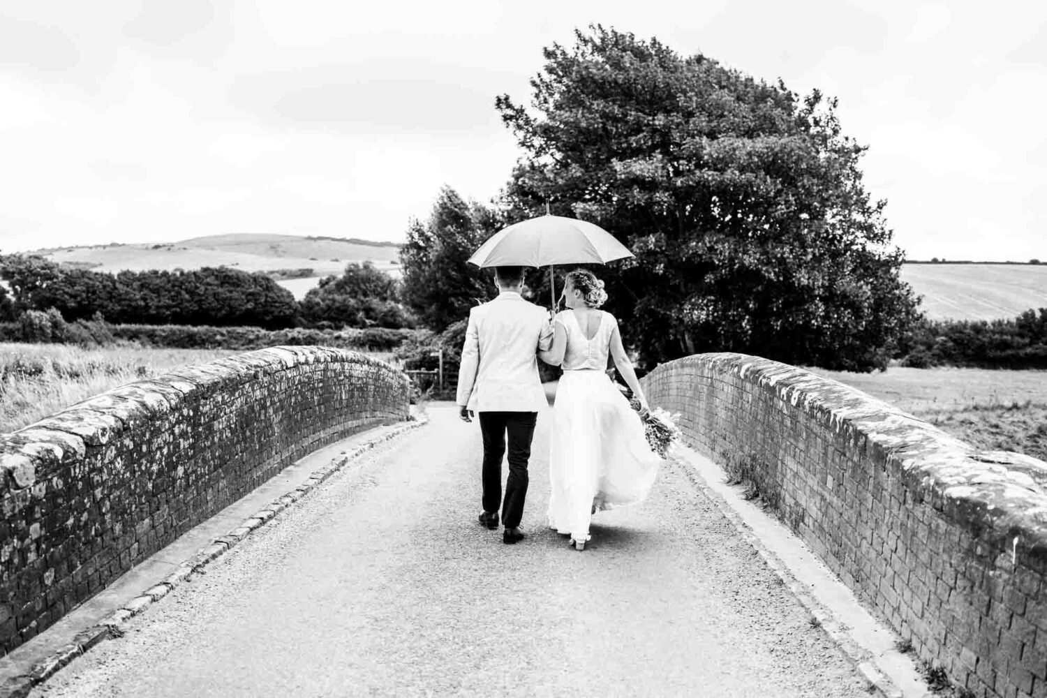 couple walking away from the camera over a bridge holding an umbrella
