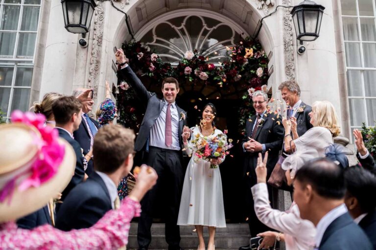 standing on steps of Chelsea old town hall groom with hand in the air confetti being thrown