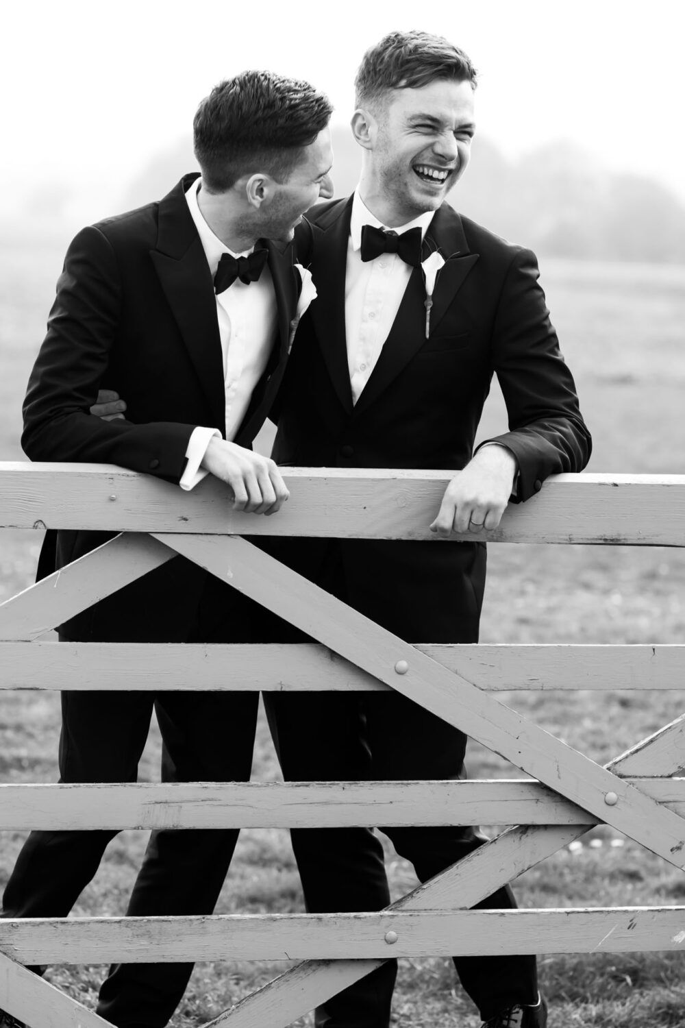 two grooms in tuxedos laughing leaning over a gate