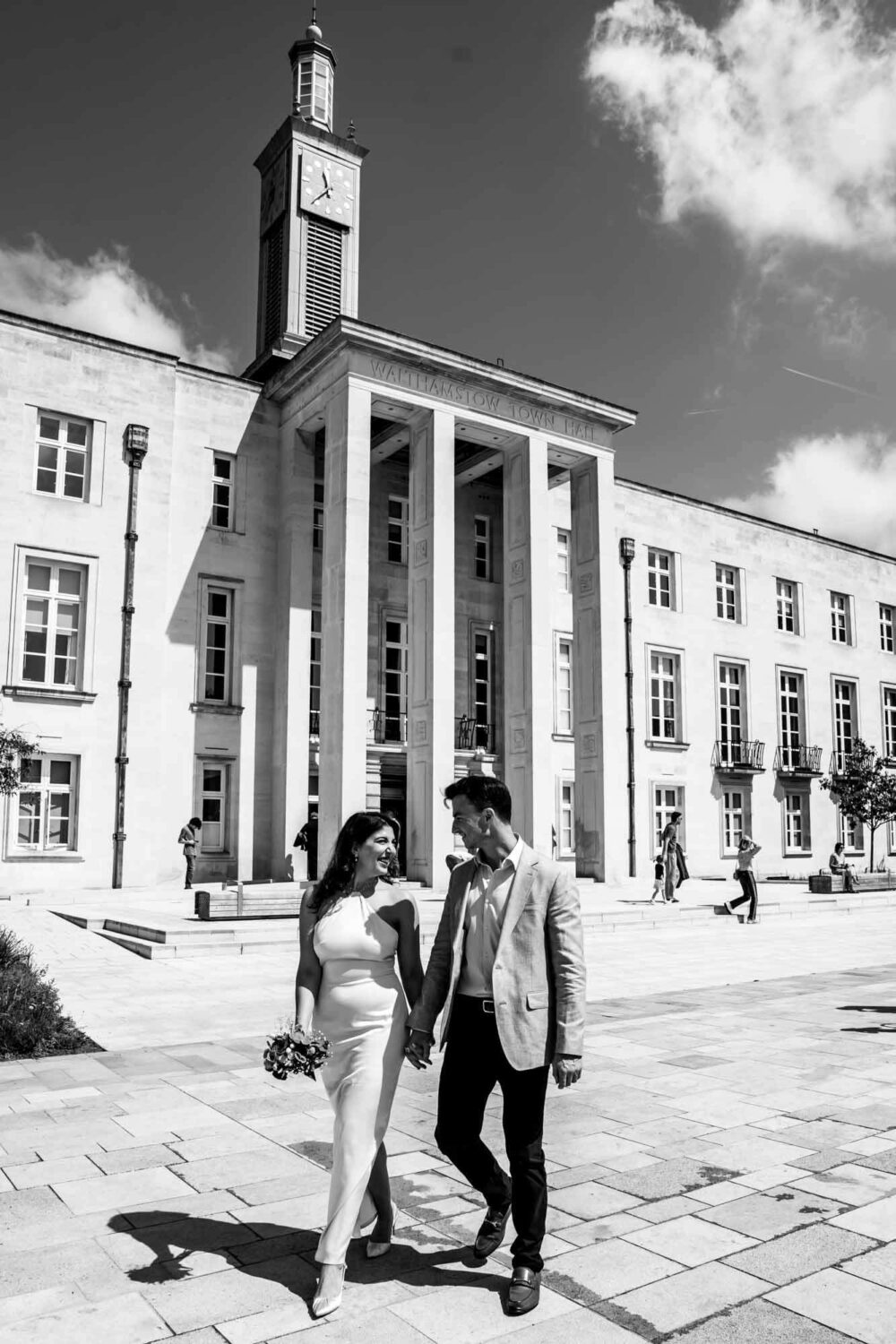 bride and groom walking hand in hand looking at each other with waltham town hall in background