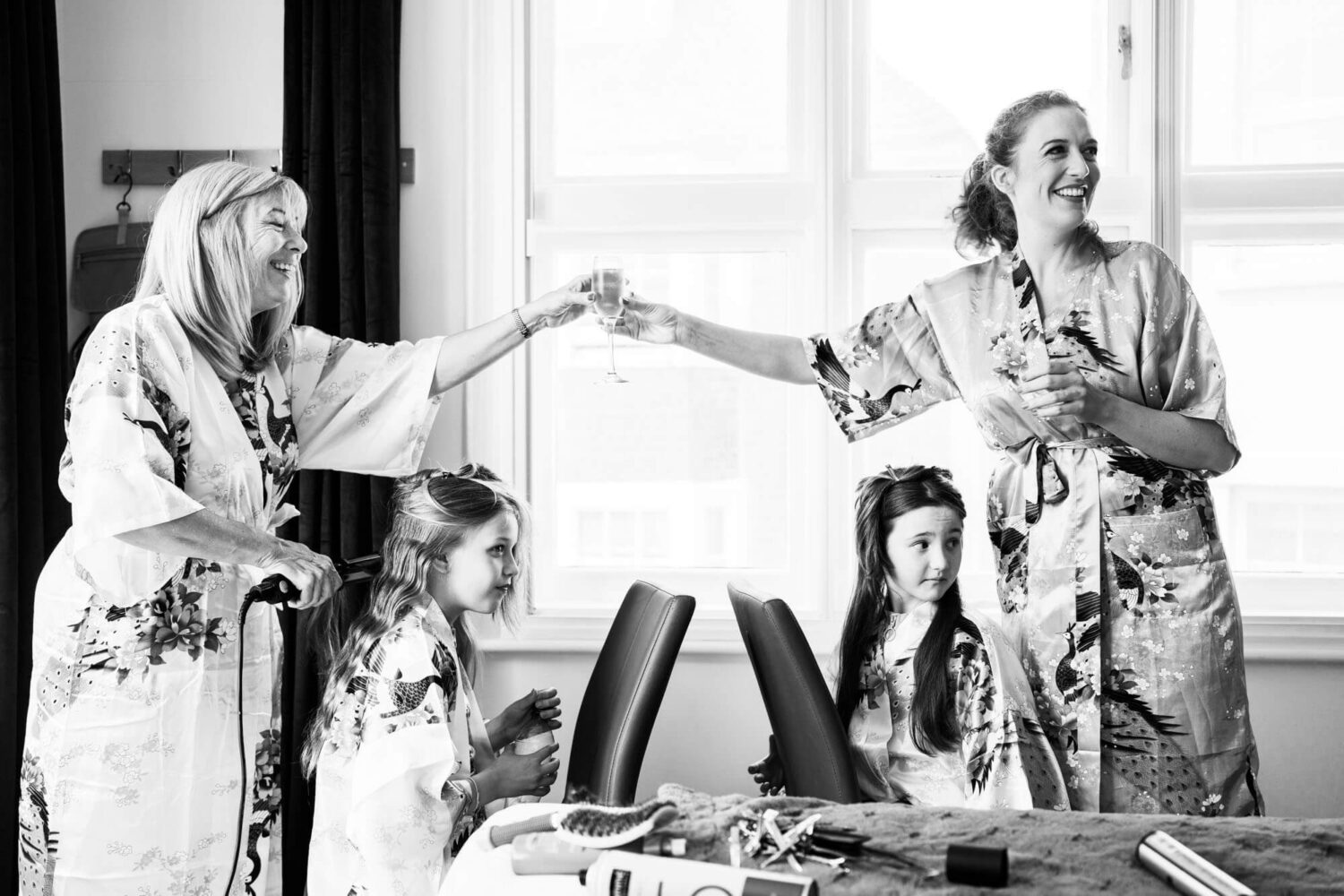mum and daughter handing over champagne with girls in chairs having hair done brighton wedding