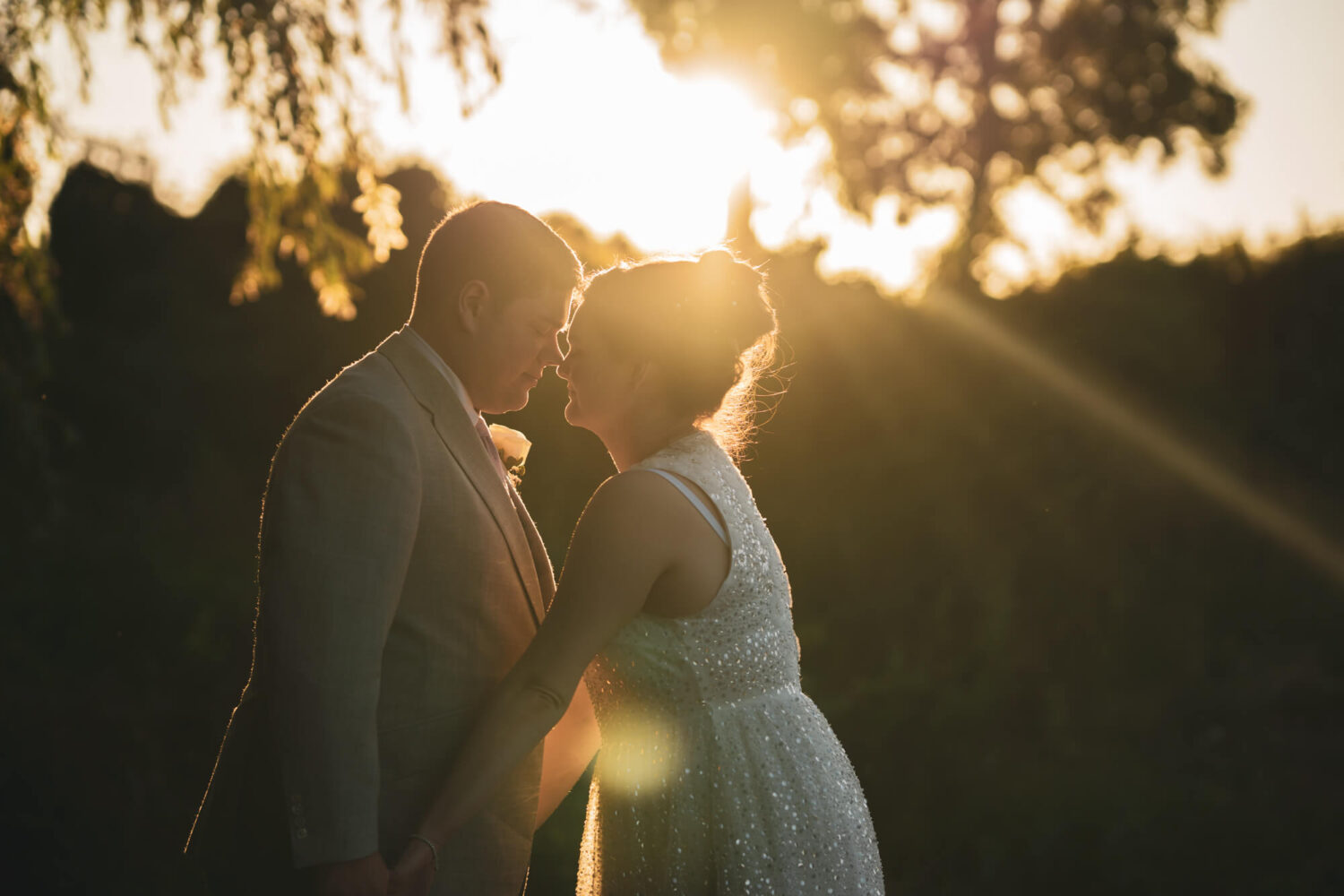 Bride and groom leaning for kiss against sunlight.