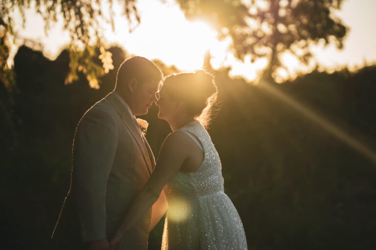 Bride and groom leaning for kiss against sunlight.