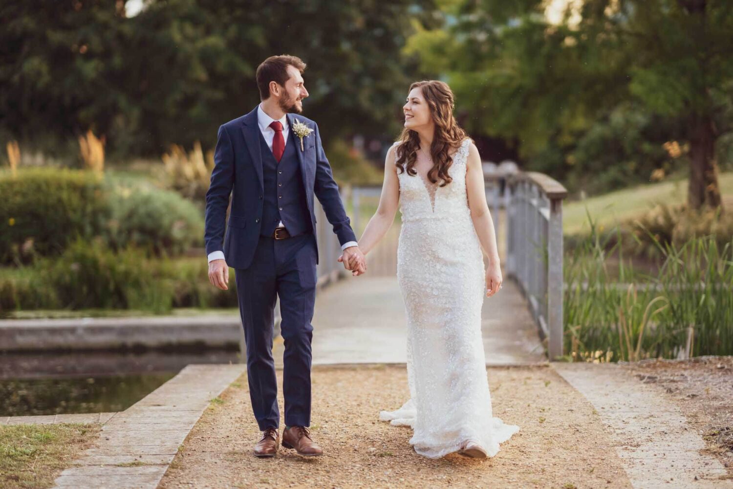 Groom and bride holding hands walking across a bridge looking at each other.