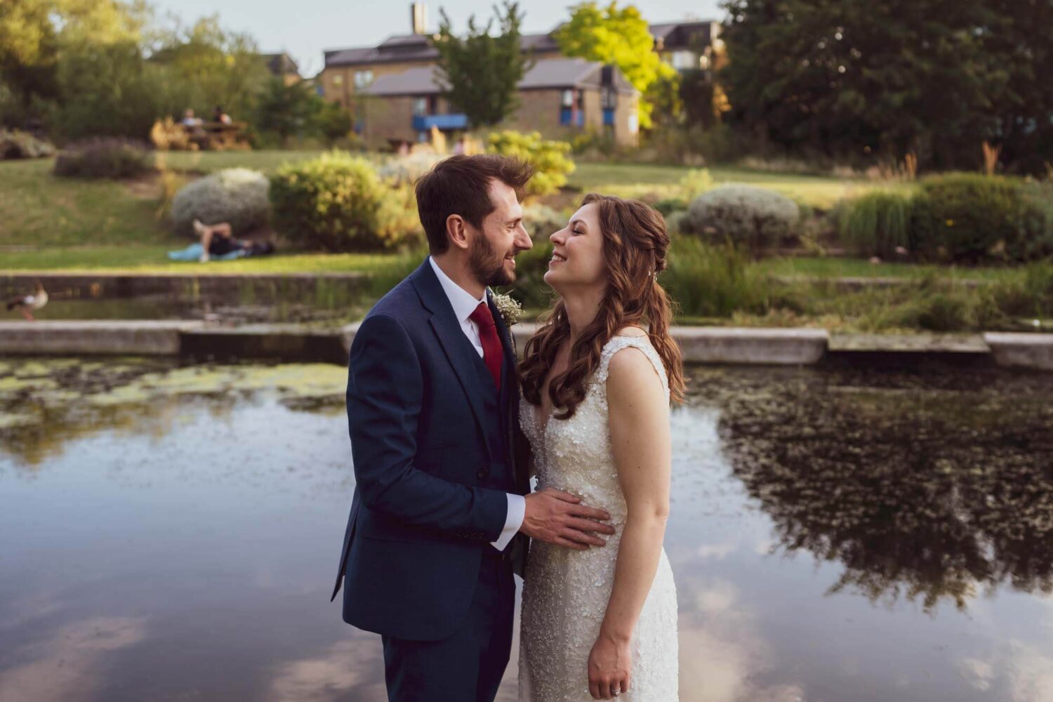 Bride and groom looking at each other arms around waist with pond in background.