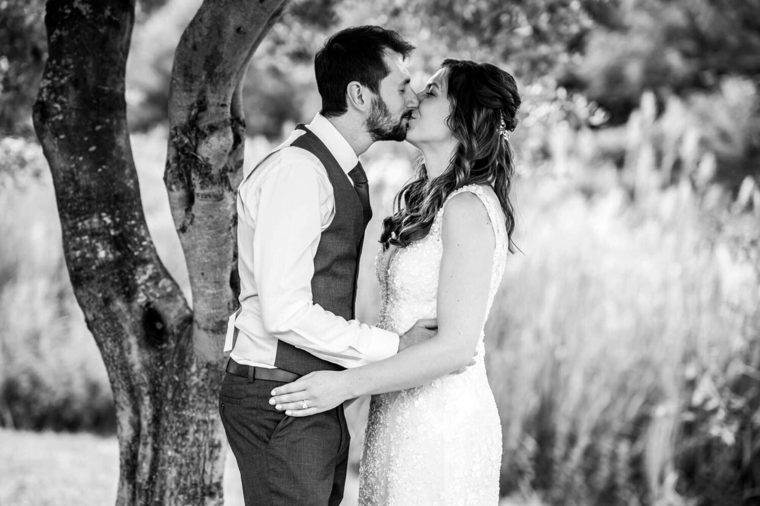 Leaning with his back to a tree, groom kissing bride with hands around her waist.