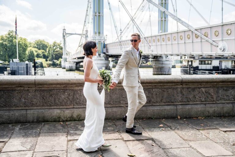 Groom taking hand of bride walking along looking back towards her by Thames Chelsea wedding.