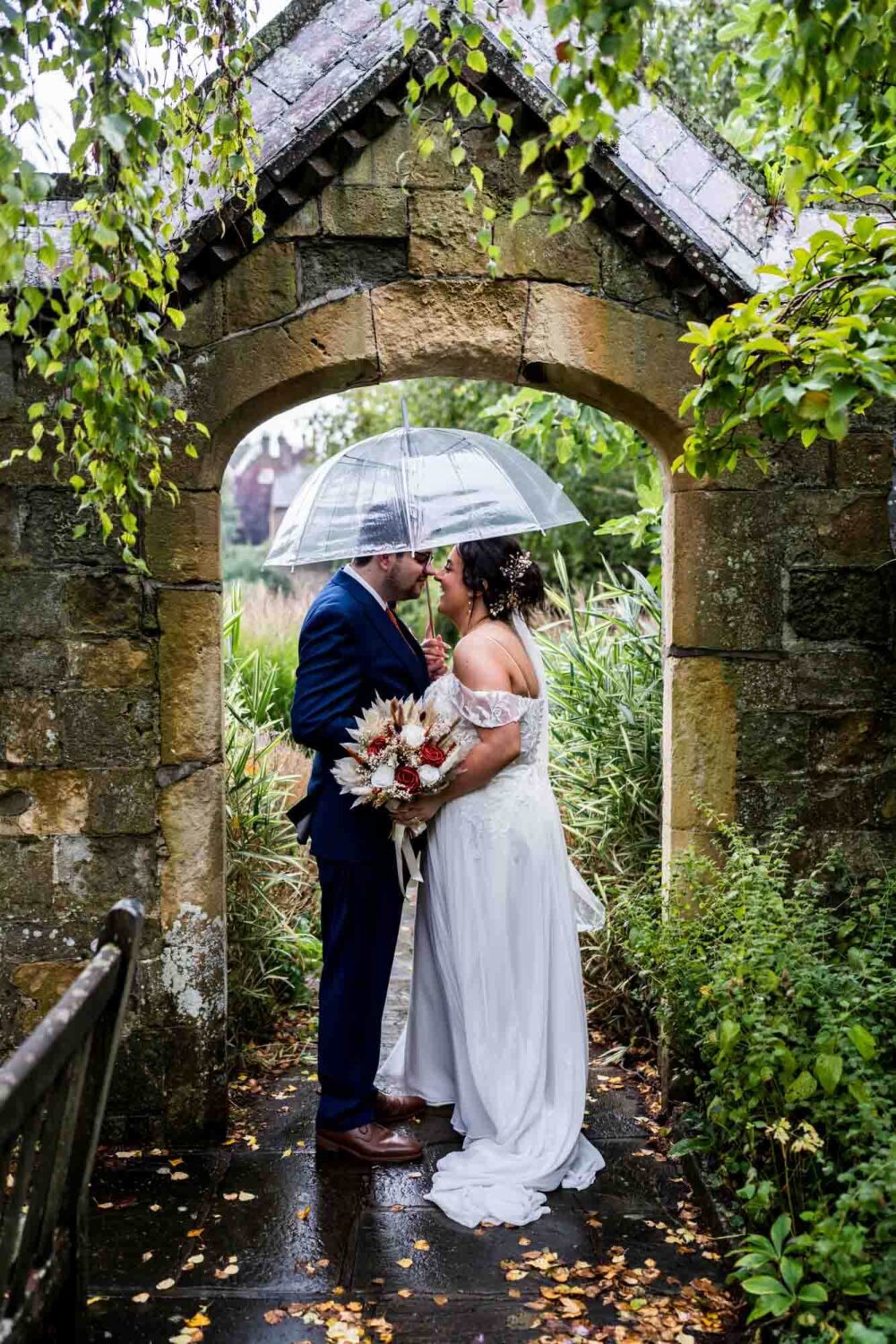 Bride and groom standing under umbrella under archway in southover grange gardens Lewes wedding.