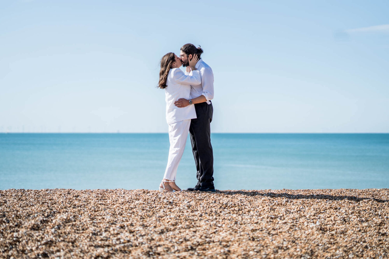 Woman bringing hands up to boyfriend's face to kiss him standing on brighton beach Brighton wedding.