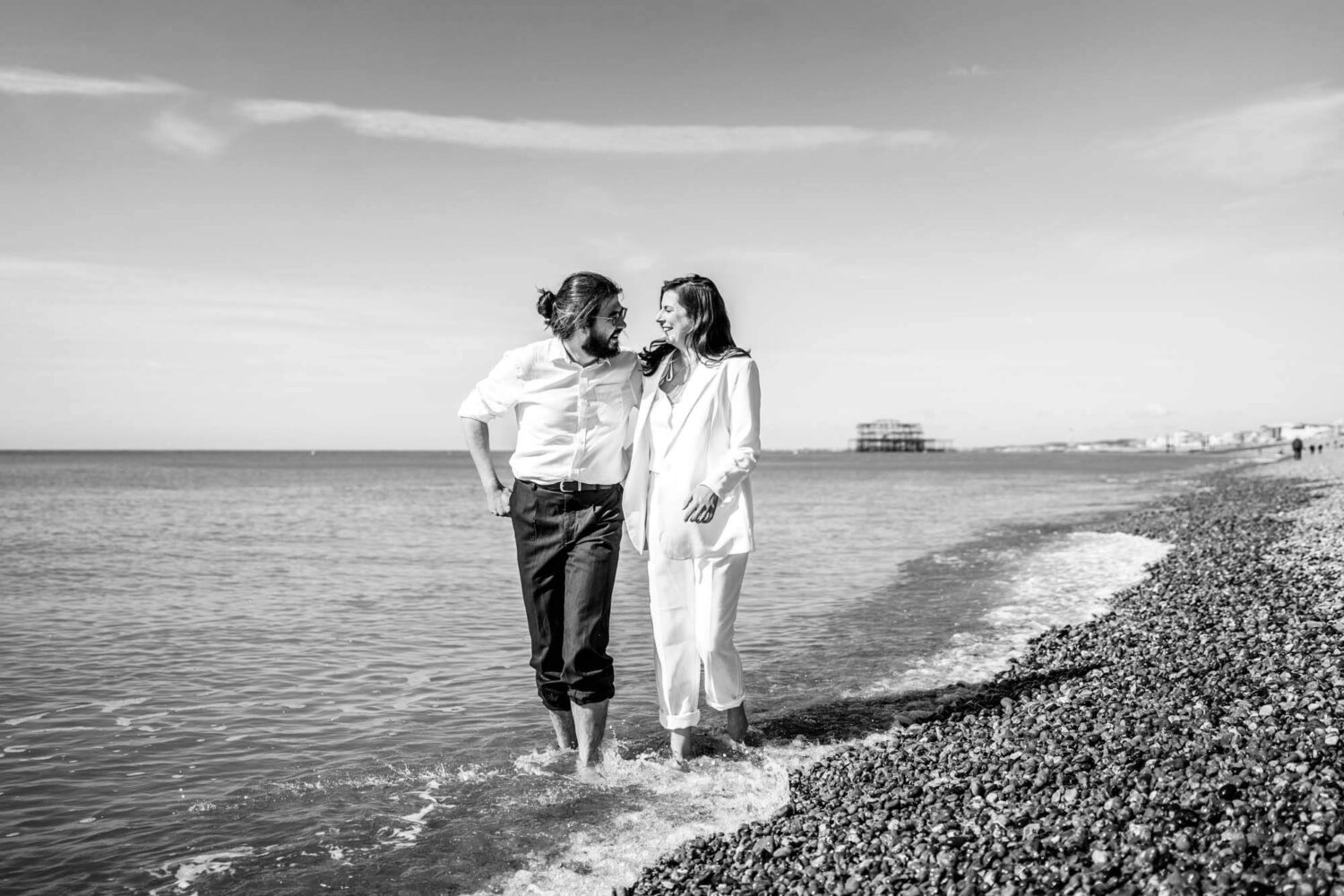Male and female couple walking through shallows of water barefoot Brighton beach.